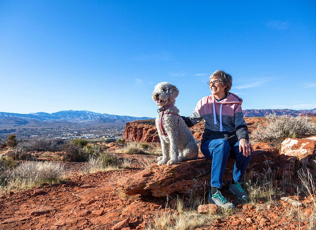 Insurance Solutions - Cheerful Senior Woman Sitting on Top of a Mountain with her Dog on a Sunny Day in the Red Rock Dessert