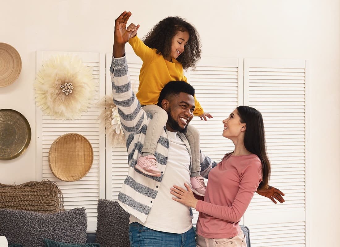 Personal Insurance - Portrait of a Cheerful Family with a Young Daughter Standing in the Living Room with the Daughter Up on her Father's Shoulders